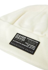 Cream-colored knitted beanie with ribbed texture; features a black fabric label with white text detailing coordinates and brand information.