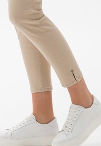 Pantalones recortados beige con una abertura lateral en el dobladillo, combinados con zapatillas blancas con cordones y suela gruesa. Textura de tela suave.