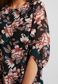 Floral top in sheer black fabric, featuring large red and white blooms. Round neckline and open sleeves with side cutouts.