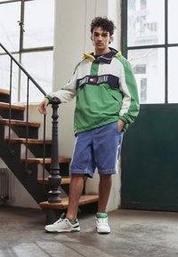 Zaļš un balts vējjakas ar tumši zilu svītru, džinsu šorti un balti sporta apavi ar zaļiem akcentiem. Modelis balstās uz kāpnēm.