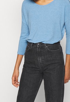 Personne portant une chemise bleu clair à manches longues rentrée dans un jean noir taille haute, debout devant un fond clair uni.