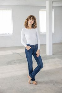 Žena s kučeravými vlasmi, bosá, oblečená v bielej blúzke s dlhým rukávom a modrých džínsoch, stojaca v jasnej prázdnej miestnosti s betónovou podlahou.