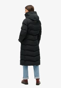 Mujer con un abrigo de plumas negro largo, con un cuello alto y capucha, jeans azul arremangados y botas marrones, de pie mirando hacia la izquierda.