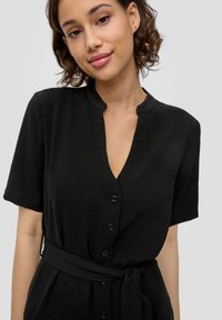 Czarna sukienka zapinana na guziki z głębokim dekoltem w kształcie litery V, krótkimi rękawami i paskiem w talii, wykonana z teksturowanej tkaniny.