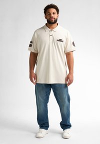 Crème polo shirt met drieknopenpat en logo-accenten op de mouwen, gecombineerd met lichtblauwe denim jeans en witte sneakers.