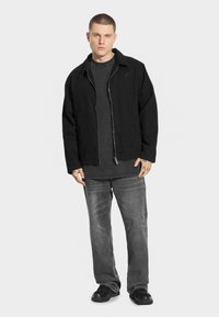 Czarna kurtka na zamek błyskawiczny noszona na ciemnoszarym podkoszulku, w zestawieniu z szarymi dżinsami. Kurtka ma klasyczny krój i subtelne logo na piersi.