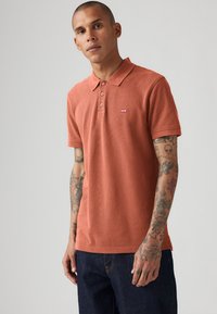 Polo de culoare ruginită cu guler ribetat, cu trei butoane, și un logo mic roșu. Mâneci scurte și purtat cu blugi de culoare albastru închis.