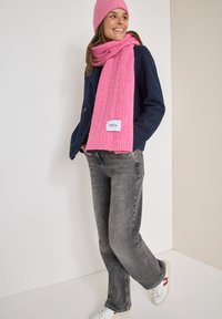 Marineblaue Fleecejacke, pinke ribbelige Strickschal, passender Beanie, graue Jeans und weiße Sneakers mit farbigen Akzenten; mit Logo-Patch auf dem Schal.