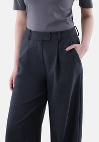 Dunkelgraue maßgeschneiderte Hose aus strukturiertem Stoff, mit Falten an der Vorderseite und seitlichen Taschen. Sie hat eine lockere Passform und Gürtelschlaufen an der Taille.
