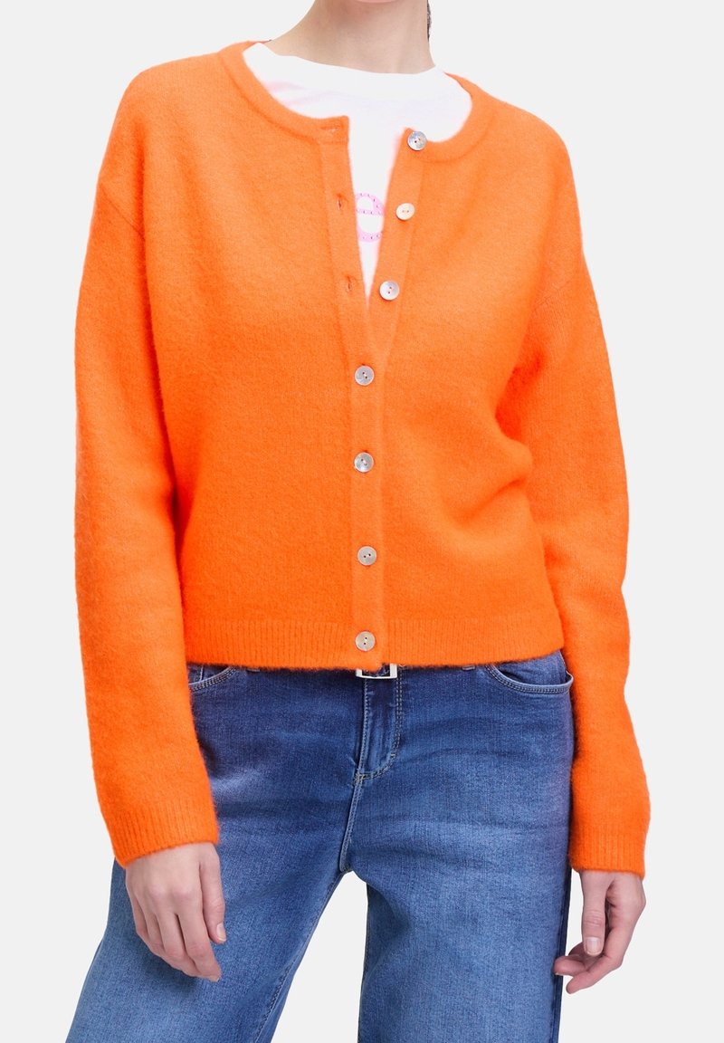 En person som bär en klar orange knapprad cardigan över en vit skjorta i kombination med blå jeans mot en vit bakgrund.