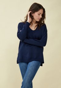 Temno moder pulover z dolgimi rokavi in V-izrezom, rebrastimi manšetami in stranskimi razpoki, v kombinaciji s svetlo modrimi jeans hlačami na nevtralni ozadju.