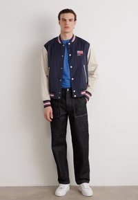 Marinblå och krämfärgad varsityjacka med ränder, blå t-shirt, mörka jeans och vita sneakers. Jackan har logotyp och fickor.