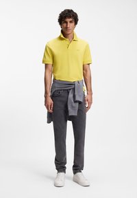 Άνδρας που στέκεται με κίτρινο πόλο πουκάμισο, γκρι τζιν, λευκά αθλητικά και γκρι φούτερ δεμένο γύρω από τη μέση του σε λευκό φόντο.