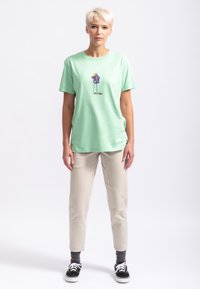 T-shirt en coton vert clair avec un design graphique au centre. Assortie à un pantalon beige et des baskets noires. Coupe simple, ajustement décontracté.