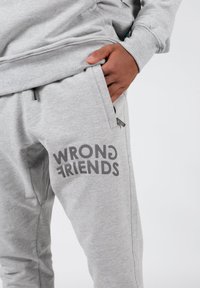 Grå sweatpants lavet af en bomuldsblanding, med teksten "WRONG FRIENDS" trykt i mørkegrå på venstre lår og sidelommer.