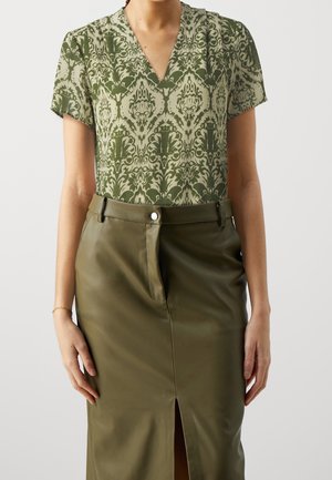 Kobieta nosząca zieloną bluzkę z krótkim rękawem, wzorzystą, z dekoltem w serek, włożoną w oliwkowozieloną skórzaną spódnicę z wysokim stanem, z rozcięciem z przodu i szlufkami na pasek.