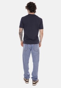 Uomo con capelli ricci, tatuaggi sul braccio sinistro, indossa una t-shirt nera semplice, jeans azzurri e scarpe da ginnastica Nike marroni, visto di spalle.