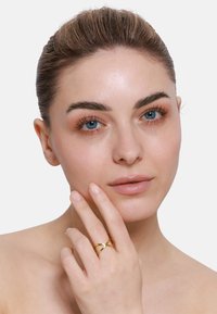 Bague croisée en ton or avec une finition polie, portée sur une main à la peau claire. Le modèle a une peau claire, des yeux bleus et un maquillage naturel.