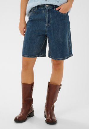 Személy kék térdig érő farmer rövidnadrágban és barna, lábszár középig érő bőrcsizmában, egyik kezét a zsebében tartva áll.