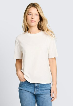 Femme blonde portant un t-shirt blanc uni à manches courtes et un jean bleu, debout avec une main dans la poche sur un fond gris clair.