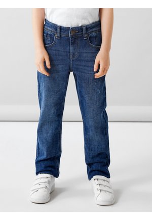 Enfant portant un jean en denim bleu et des baskets blanches, debout devant un fond gris uni.