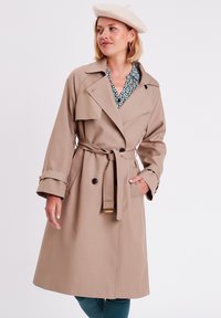 Gabardina beige con cinturón, grandes solapas, botones de doble pecho y mangas largas. Combinada con una camisa con estampado y una boina.