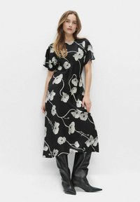 Kobieta w czarnej sukience midi w białe kwiatowe wzory oraz bufiastymi rękawami, zestawiona z czarnymi skórzanymi kozakami do kolan.