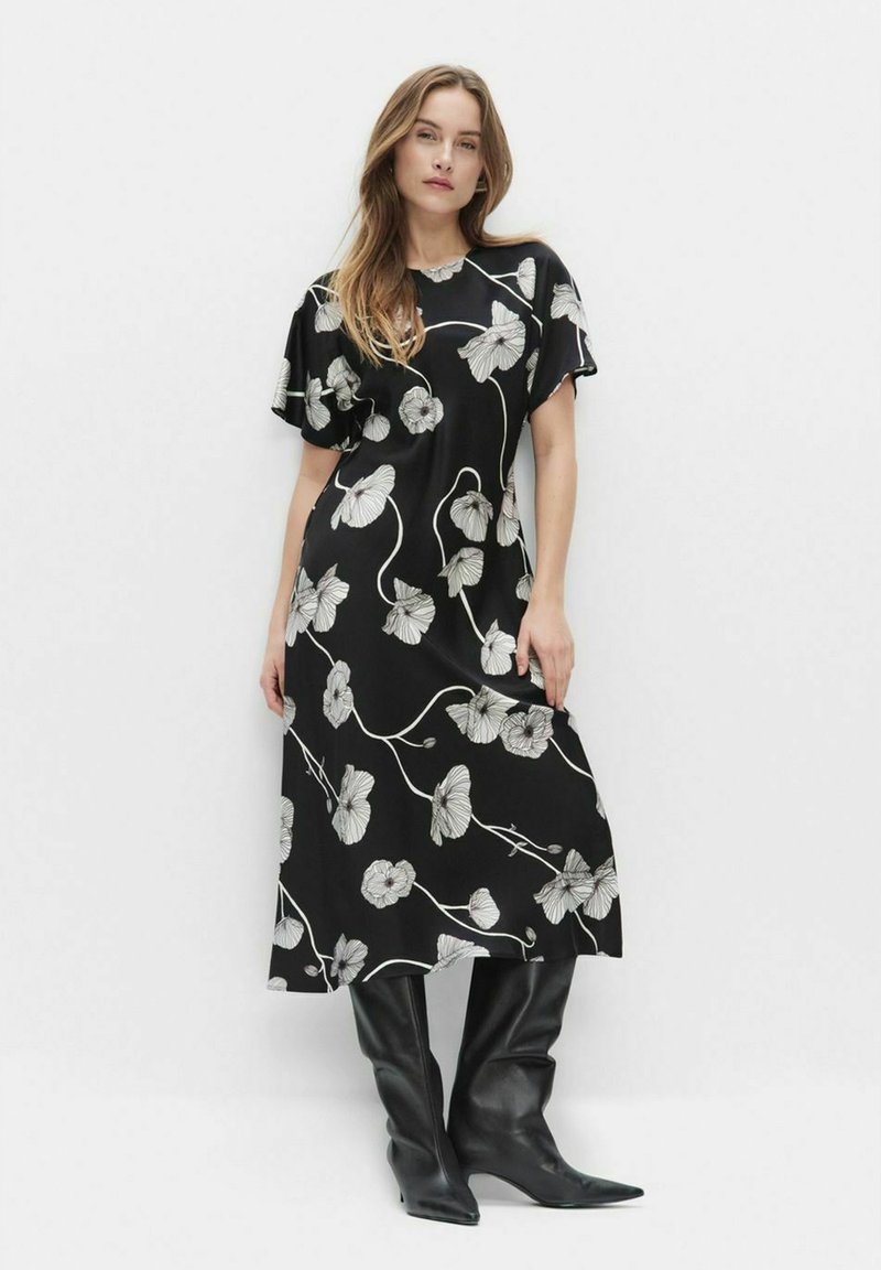 Kobieta w czarnej sukience midi w białe kwiatowe wzory oraz bufiastymi rękawami, zestawiona z czarnymi skórzanymi kozakami do kolan.