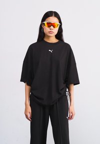 Svart oversized T-skjorte med hvit logo, side snorer, kombinert med svarte bukser. Modellen har refleksjoner solbriller, og viser en sporty stil.