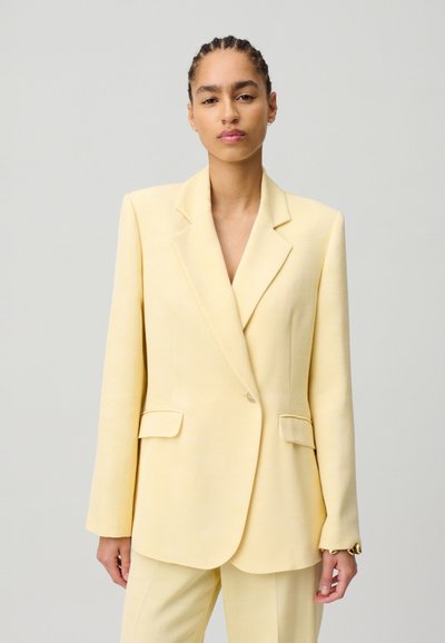 Blazer giallo con una vestibilità su misura, rever a punta e tasche frontali. Realizzato in un tessuto strutturato con una consistenza liscia. Dettagli hardware minimi.