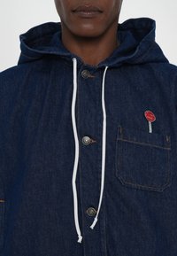 Mørkeblå denim hoodiejakke med hvid snøre, sølvknapper og en rød lollipop brodering på brystlommen.