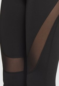 Nærbillede af sorte leggins med gennemsigtige mesh-paneler på lår og knæ, som fremhæver tekstur og syninger i stoffet.