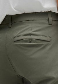 Oliwkowe spodnie o gładkiej fakturze, wyposażone w tylną kieszeń z zapięciem na guzik oraz szlufkami na pasek wzdłuż pasa.