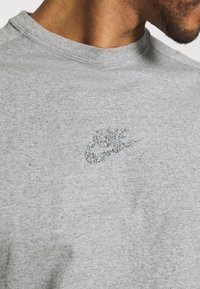 Šedé bavlněné tričko s třpytivým stříbrným logem Nike na hrudi, se standardním kulatým výstřihem a krátkým rukávem.