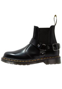 Bottine en cuir noir avec panneau élastique sur le côté, sangle et détails de boucle, surpiqûres jaunes et un tirette en boucle sur une semelle translucide.