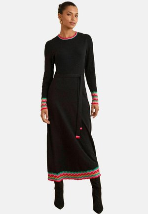 Schwarzes Strickkleid mit langen Ärmeln, das mit einem mehrfarbigen Zickzackbesatz in Rot und Grün am Halsausschnitt und an den Ärmeln sowie einem passenden Taillengürtel versehen ist.