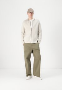 Ljusbeige zip-up hoodie, olivgröna byxor, vita sneakers. En enkel design med en avslappnad passform, tillverkad av mjukt material.