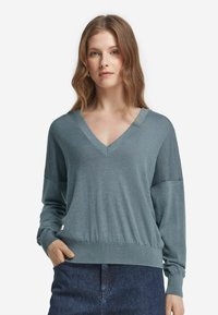 Kvinna med vågigt ljusbrunt hår som bär en blå V-ringad långärmad tröja och mörkblå jeans, stående med en hand i fickan.