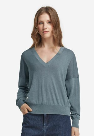 Frau mit welligem, hellbraunem Haar, die einen blauen V-Ausschnitt Langarmpullover und dunkelblaue Jeans trägt und mit einer Hand in ihrer Tasche steht.