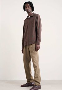 Geruit overhemd in bruin, groen en rood; klassieke kraag; enkele borstzak. Beigekleurige broek met een rechte snit en bruine schoenen.