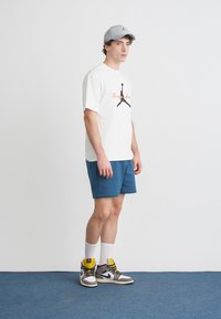 T-shirt blanc avec un motif graphique noir, short bleu, casquette grise, chaussettes blanches et baskets hautes multicolores, sur un fond neutre.