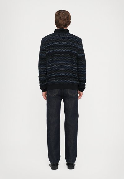 Melnas adītas kardigānas ar ziliem zigzag rakstiem, ribotām apkaklēm un piedurknēm. Apvienojums ar tumšas džinsām, skatīts no aizmugures.