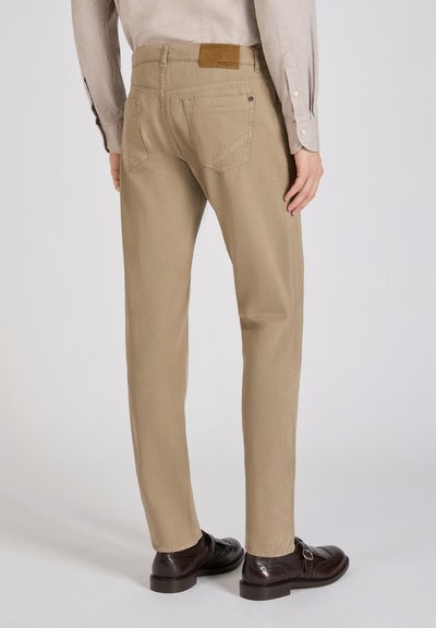 Pantalones beige de corte recto, con bolsillos traseros y una etiqueta de cuero marrón en la cintura. Combinados con zapatos de vestir oscuros.