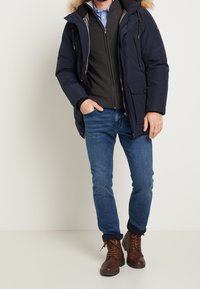 Blå puffjacka med fuskpäls på huvan, grå tröja och blå jeans, kombinerad med bruna snörstövlar, stående mot en neutral bakgrund.