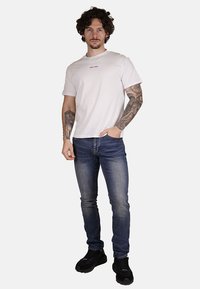 Maglietta di cotone bianca con un piccolo logo multicolore, abbinata a jeans in denim blu e scarpe sportive nere. Il modello sta in posizione eretta.