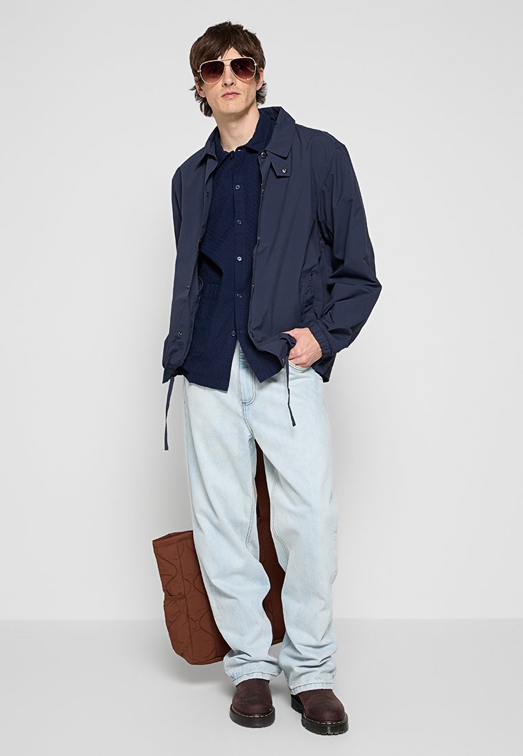 Marineblauer Windbreaker über einem strukturierten blauen Hemd, hellblaue lockere Jeans, braune Slipper und große Sonnenbrille. Braune gesteppte Tasche.