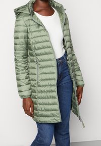 Olivengrøn pufferjakke med hætte, med vandret syninger, lynlåslommer og et mellemlangt design. Båret over en hvid bluse og denim.