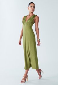 Olijfgroene, mouwloze maxi-jurk met een diepe V-hals, geplooide taille, zachte textuur en een vloeiende silhouet. Gecombineerd met witte sandalen met bandjes.