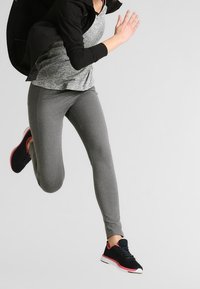 Persona corriendo con mallas grises, chaqueta negra y zapatillas negras con suelas rosas, sobre un fondo liso claro.