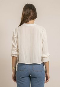 Blouse blanche texturée avec de longues manches bouffantes et un ourlet court, associée à un jean en denim bleu. Le tissu présente de légères lignes verticales.